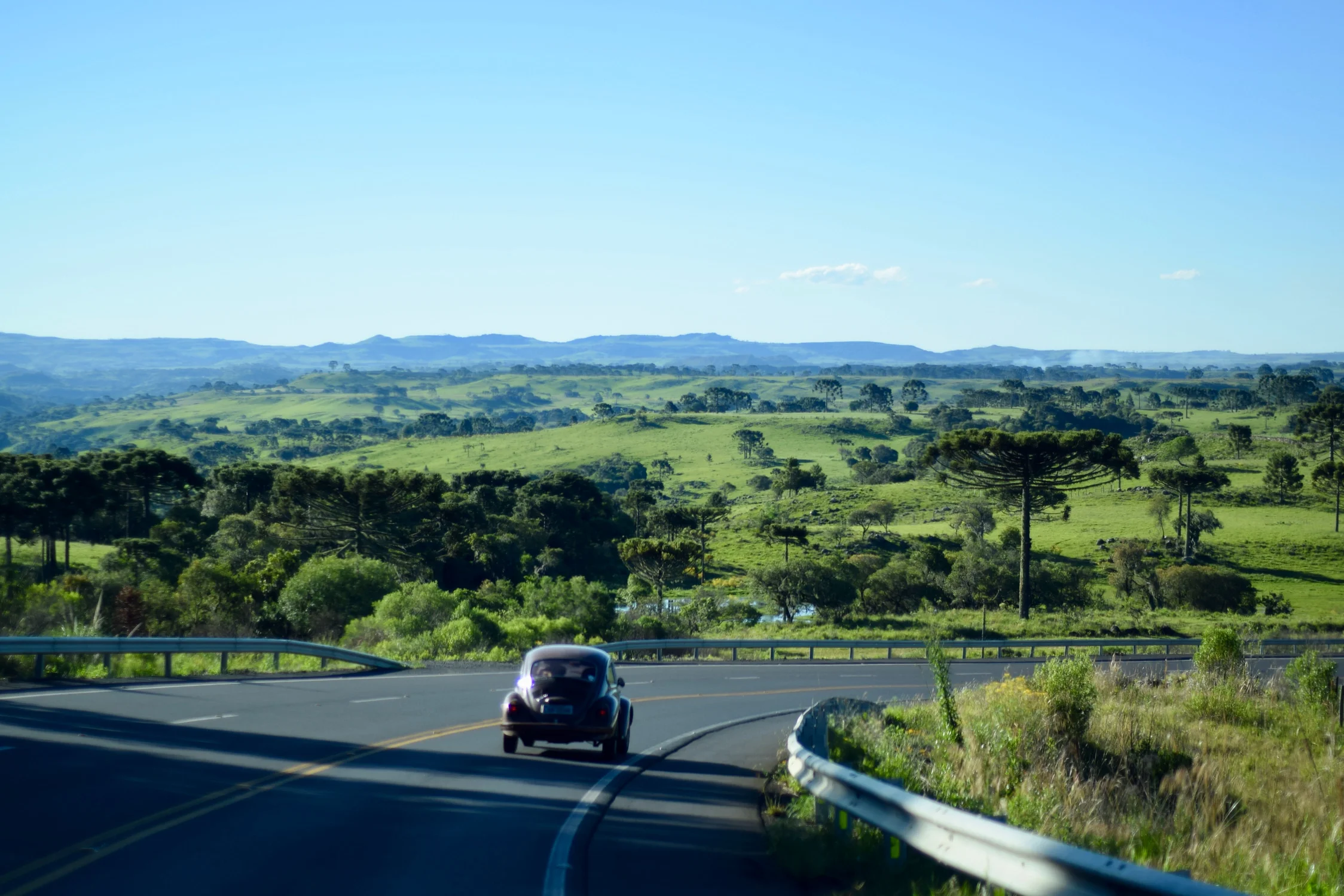 Seguro Automovel: VW Fusca andando na rodovia, com uma bela paisagem montanhosa e bem arborizada.