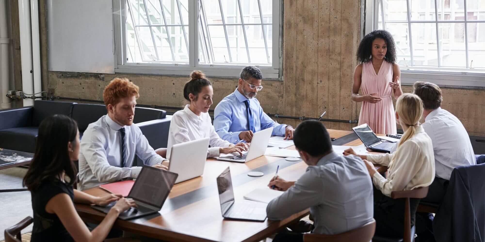 Seguro de Vida Empresarial: Uma mulher em pé, fazendo uma reunião na frente de uma mesa, com mais funcionarios sentados e notebooks em cima da mesa.